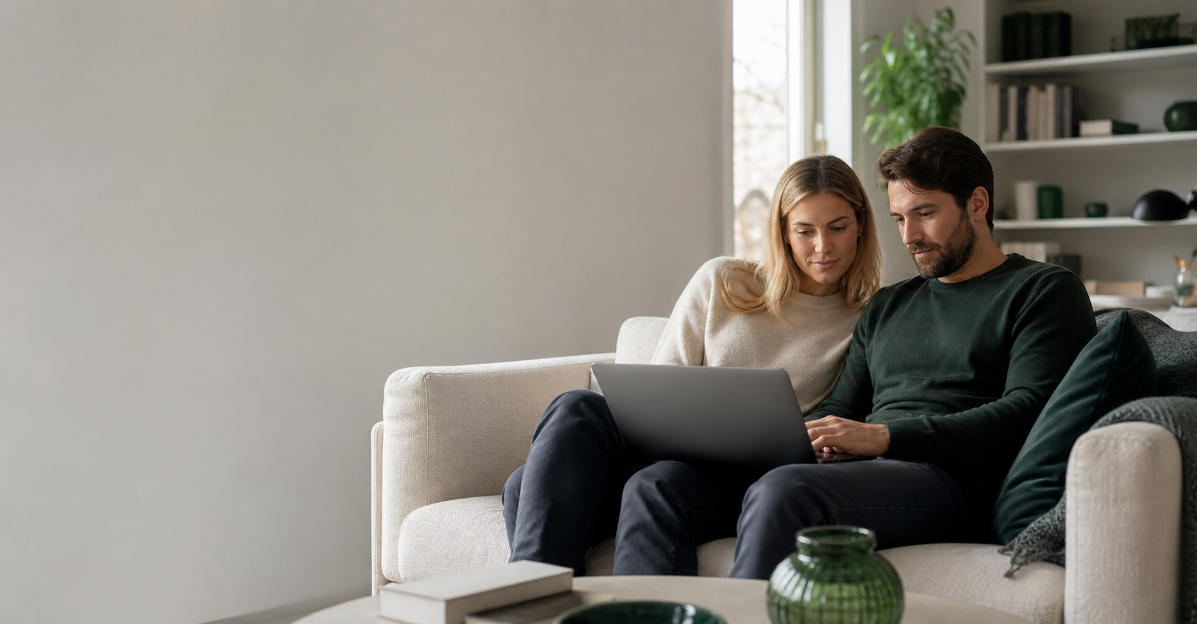 Ein junges Paar sitzt auf dem Sofa im Wohnzimmer und sieht sich gemeinsam am Laptop seine Baufinanzierung an.